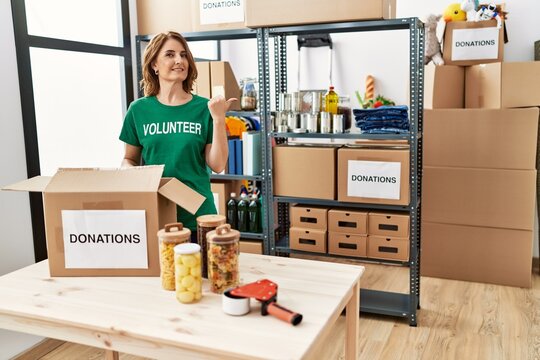 Middle age woman wearing volunteer t shirt at donations stand pointing thumb up to the side smiling happy with open mouth