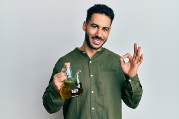 Young hispanic man holding olive oil can doing ok sign with fingers, smiling friendly gesturing...