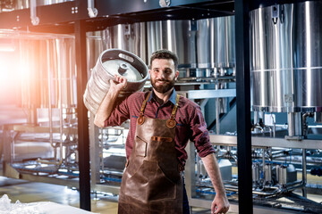 Young male brewer in leather apron holds barrel with craft beer at modern brewery factory