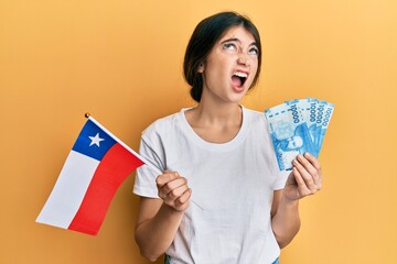 Young caucasian woman holding chile flag and chilean pesos banknotes angry and mad screaming frustrated and furious, shouting with anger looking up.