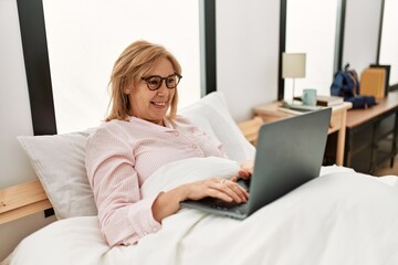 Obraz premium Middle age blonde woman smiling happy using laptop lying on the bed at home.