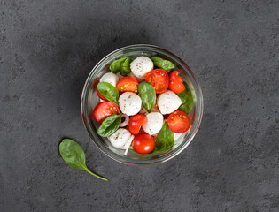 Caprese salad of tomatoes, onions, spinach and mozzarella in a glass bowl on a dark gray background top view