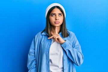 Young latin woman wearing cute wool cap thinking concentrated about doubt with finger on chin and...