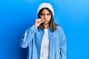 Young latin woman wearing cute wool cap mouth and lips shut as zip with fingers. secret and silent,...