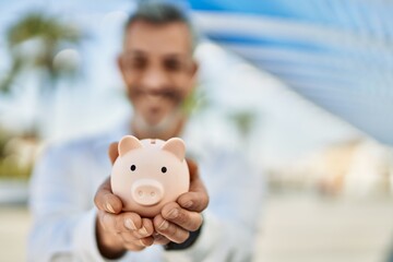 Middle age grey-haired man smiling happy holding piggy bank at the city.