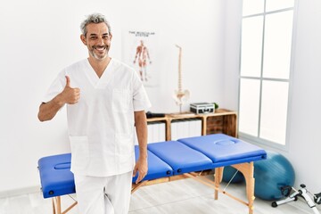 Middle age hispanic therapist man working at pain recovery clinic doing happy thumbs up gesture with hand. approving expression looking at the camera showing success.