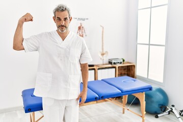 Middle age hispanic therapist man working at pain recovery clinic strong person showing arm muscle, confident and proud of power
