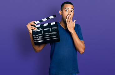 Young african american man holding video film clapboard covering mouth with hand, shocked and...