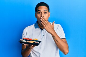Young handsome hispanic man showing a plate of sushi covering mouth with hand, shocked and afraid...