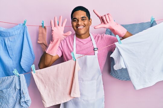 Young handsome hispanic man wearing cleaner apron holding clothes on clothesline showing and pointing up with fingers number seven while smiling confident and happy.
