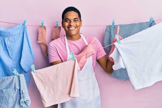 Young handsome hispanic man wearing cleaner apron holding clothes on clothesline smiling and looking at the camera pointing with two hands and fingers to the side.