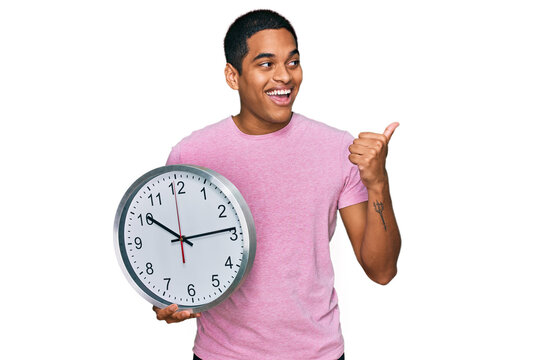 Young handsome hispanic man holding big clock pointing thumb up to the side smiling happy with open mouth
