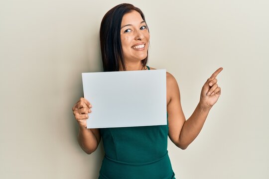 Young latin woman holding blank empty banner smiling happy pointing with hand and finger to the side