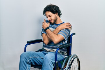 Naklejka premium Handsome hispanic man sitting on wheelchair hugging oneself happy and positive, smiling confident. self love and self care