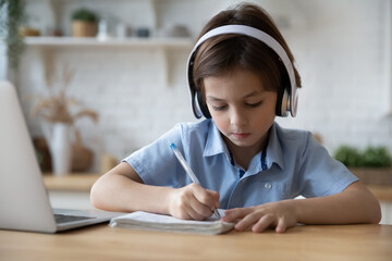 Naklejka premium Concentrated little child boy in headphones writing notes in copybook, studying on online school lessons remotely from home. Happy cute kid studying distantly, learning foreign language, doing tasks.