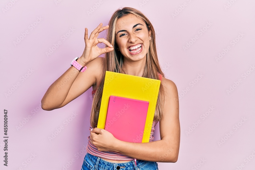 Canvas Prints Beautiful hispanic woman holding books doing ok sign with fingers, smiling friendly gesturing excellent symbol