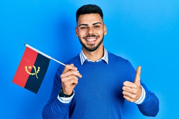 Young hispanic man with beard holding angola flag smiling happy and positive, thumb up doing excellent and approval sign