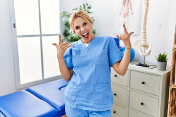 Beautiful blonde physiotherapist woman working at pain recovery clinic shouting with crazy...