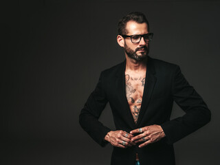 Portrait of handsome confident stylish hipster lambersexual model. Sexy modern man dressed in black jacket. Naked torso with tattoos.Fashion male posing in studio on dark background. In spectacles
