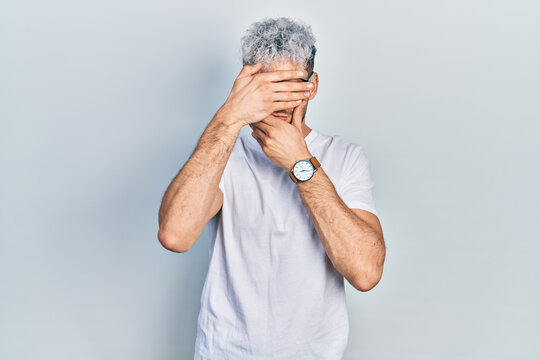Young hispanic man with modern dyed hair wearing white t shirt and glasses covering eyes and mouth with hands, surprised and shocked. hiding emotion