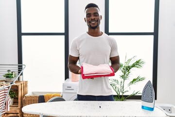 Young african man holding folded laundry after ironing smiling with a happy and cool smile on face. showing teeth.