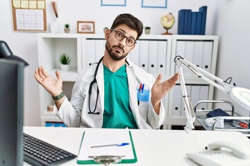 Young man with beard wearing doctor uniform and stethoscope at the clinic clueless and confused expression with arms and hands raised. doubt concept.