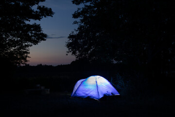 A bright blue glowing tent in a tourist parking lot against the background of dark silhouettes of trees. Summer night with clear blue starry sky.