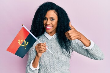 Middle age african american woman holding angola flag smiling happy and positive, thumb up doing excellent and approval sign