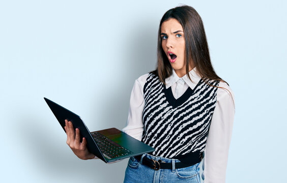 Young brunette teenager working using computer laptop scared and amazed with open mouth for surprise, disbelief face