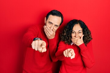 Middle age couple of hispanic woman and man hugging and standing together laughing at you, pointing finger to the camera with hand over mouth, shame expression