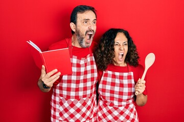 Middle age couple of hispanic woman and man wearing professional apron reading cooking recipe book angry and mad screaming frustrated and furious, shouting with anger. rage and aggressive concept.