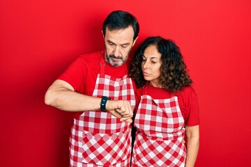Middle age couple of hispanic woman and man wearing cook apron checking the time on wrist watch, relaxed and confident