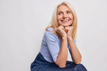 Indoor shot of dreamy middle aged blonde woman keeps hands under chin recalls pleasant memory in mind smiles broadly sits against grey background blank empty space for your advertisement or text - obrazy, fototapety, plakaty