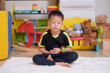 Little Asian kid with eyes closed, barefoot practices yoga and meditating to relieve negative...