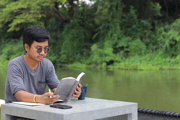 Asian man with black hair and glasses is taking a weekend vacation in nature by the river. reading a book in hopes of bringing his knowledge to become the author of his dream book happily and relax
