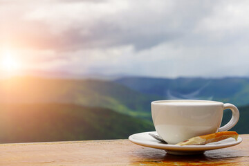 A white cup of hot espresso coffee mugs placed on a wooden floor with morning fog and mountains with sunlight background,coffee morning