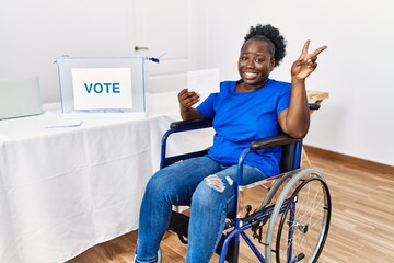 Young african woman sitting on wheelchair voting putting envelop in ballot box smiling looking to the camera showing fingers doing victory sign. number two.