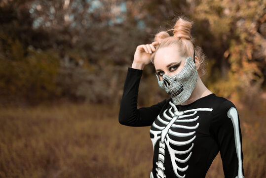 A girl with a scary make-up in a skeleton costume, a mask with rhinestones in a gloomy autumn forest. Stands terribly, looking at the camera. Portrait. Halloween, autumn holiday concept 
