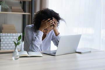 Professional burnout. Depressed upset mixed race female sit at workplace by computer hug head feel unmotivated sick lack of sleep. Desperate young black woman having problems at work missed deadline - obrazy, fototapety, plakaty