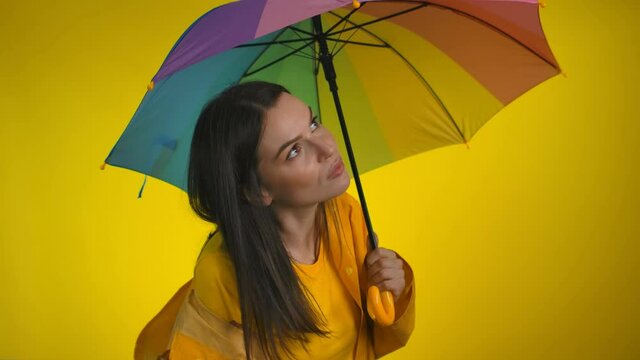 A happy woman in yellow raincoat with rainbow umbrella is checking for the rain