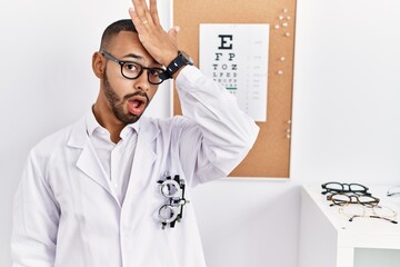 African american optician man standing by eyesight test surprised with hand on head for mistake, remember error. forgot, bad memory concept.