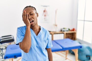 Black woman with braids working at pain recovery clinic yawning tired covering half face, eye and mouth with hand. face hurts in pain.