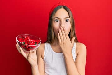 Beautiful brunette little girl holding red pepper covering mouth with hand, shocked and afraid for...