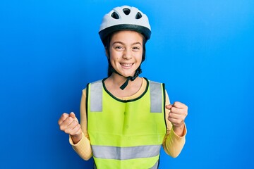 Beautiful brunette little girl wearing bike helmet and reflective vest very happy and excited doing winner gesture with arms raised, smiling and screaming for success. celebration concept.