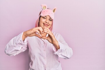 Hispanic woman with pink hair wearing sleep mask and pajama smiling in love doing heart symbol shape with hands. romantic concept.