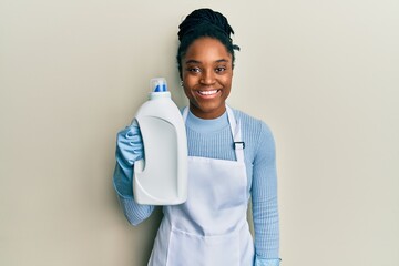 African american woman with braided hair holding detergent bottle looking positive and happy standing and smiling with a confident smile showing teeth