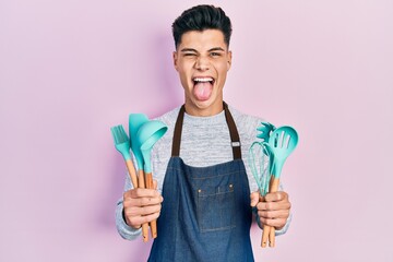 Young hispanic man wearing professional baker apron holding cooking tools sticking tongue out happy...