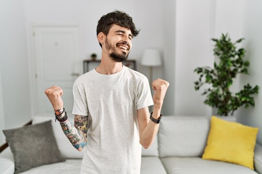 Hispanic man with beard at the living room at home celebrating surprised and amazed for success with arms raised and eyes closed. winner concept.