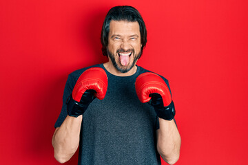 Middle age caucasian man using boxing gloves sticking tongue out happy with funny expression.