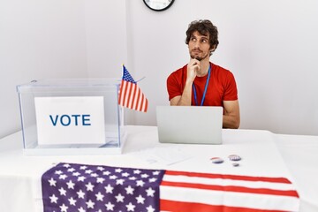 Young hispanic man at political election sitting by ballot with hand on chin thinking about question, pensive expression. smiling with thoughtful face. doubt concept.